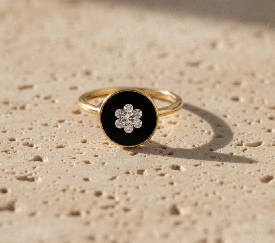 gold ring with a round black enamel top featuring a sparkling flower design made of clear stones, photographed on a textured beige surface in natural sunlight.